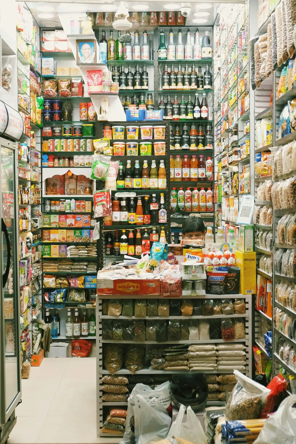 Pequeña tienda de abarrotes llena de productos empacados, con el tendero detrás del mostrador